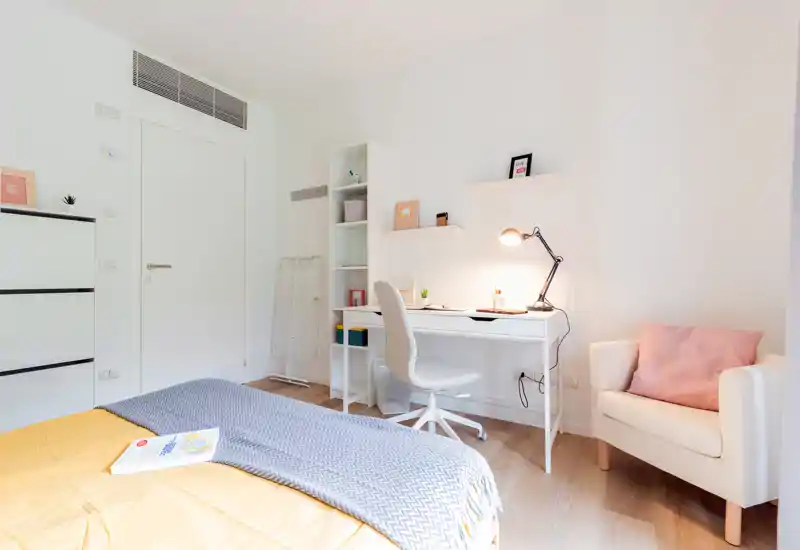 Dormitorio moderno y luminoso con la cama bien hecha, manta acogedora y escritorio con lámpara — ideal para una sola persona o estilo estudio.