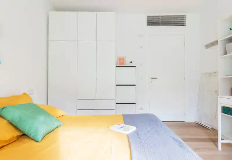Dormitorio moderno y luminoso con cama bien hecha en ropa de cama amarilla, cojines verdes y armarios blancos minimalistas que crean un ambiente fresco y acogedor.