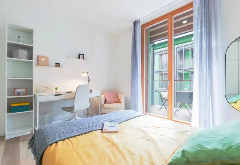 Dormitorio moderno y luminoso con cama cómoda, textiles suaves y zona de escritorio. Grandes puertas de cristal se abren a un balcón con abundante luz natural.