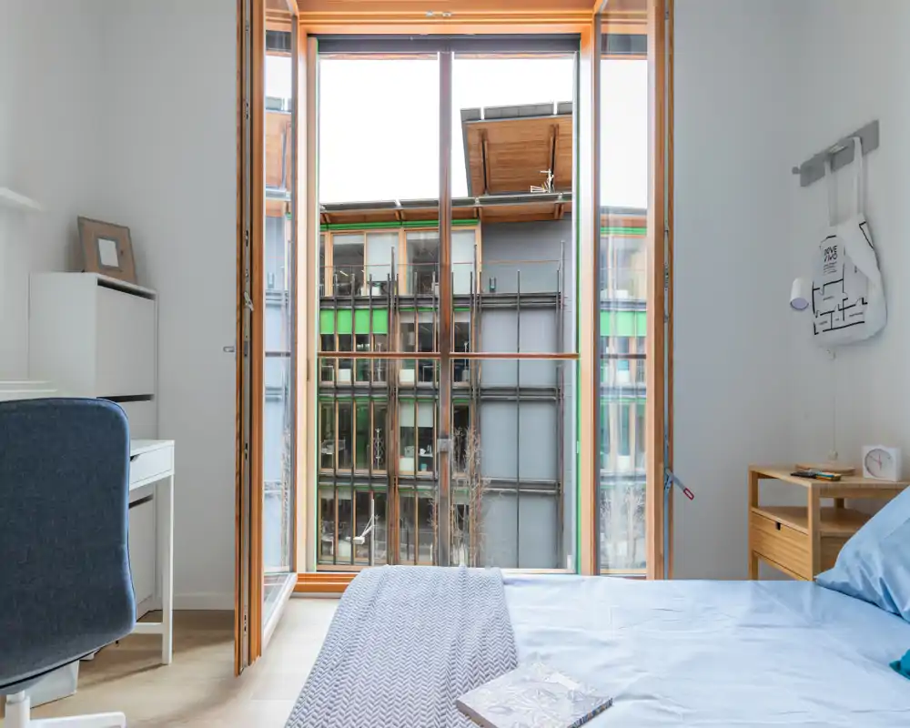Camera da letto moderna e luminosa con ampia finestra a tutta parete, letto ben rifatto e comodini essenziali — perfetta per affitti in città con molta luce naturale.