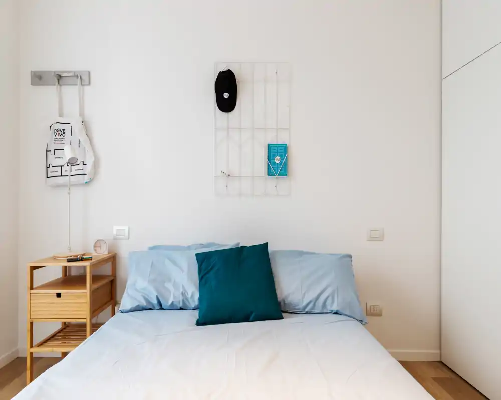 Camera da letto luminosa e minimalista con letto matrimoniale ben rifatto, biancheria azzurro chiaro e cuscino accent color verde petrolio. Un piccolo comodino in legno e ganci a muro completano l'arredo semplice e arioso, perfetto per annunci.