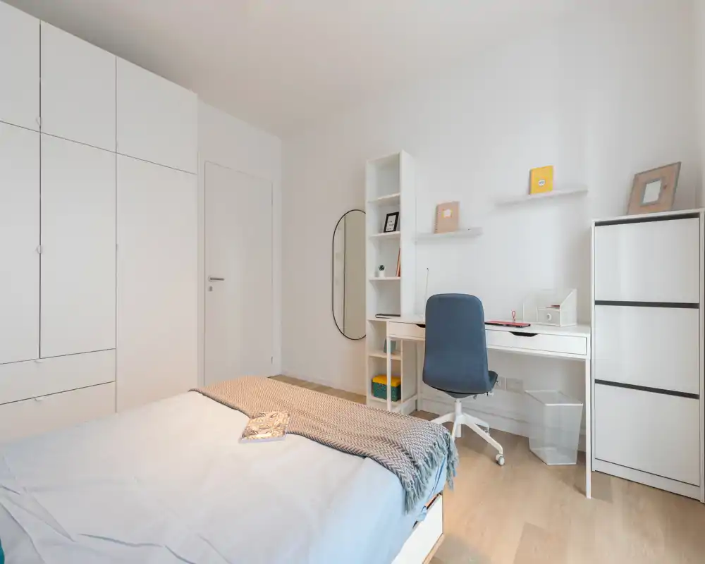 Camera da letto luminosa e moderna con letto ben fatto, armadi a muro e un angolo studio compatto. Pavimento in legno chiaro e arredi minimali creano uno spazio pulito e accogliente, perfetto per affitti brevi.