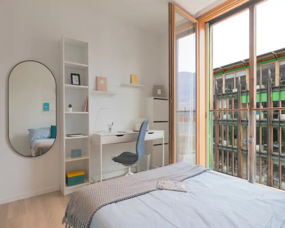 Camera da letto moderna e luminosa con letto rifatto, scrivania per lo studio e ampia finestra dal pavimento al soffitto che offre molta luce naturale e vista sulla città.