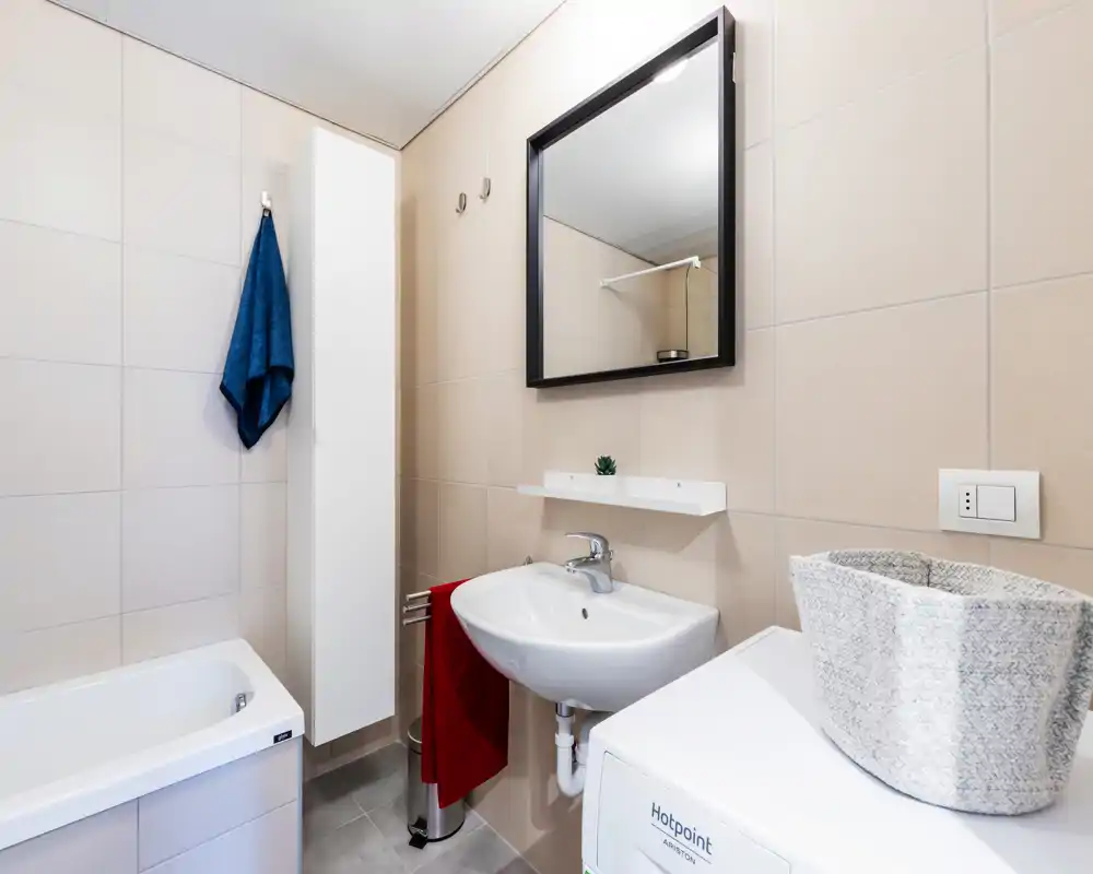 Bagno moderno e luminoso con lavabo, specchio, vasca e lavatrice; piastrelle neutre e accessori curati creano uno spazio pulito e funzionale.