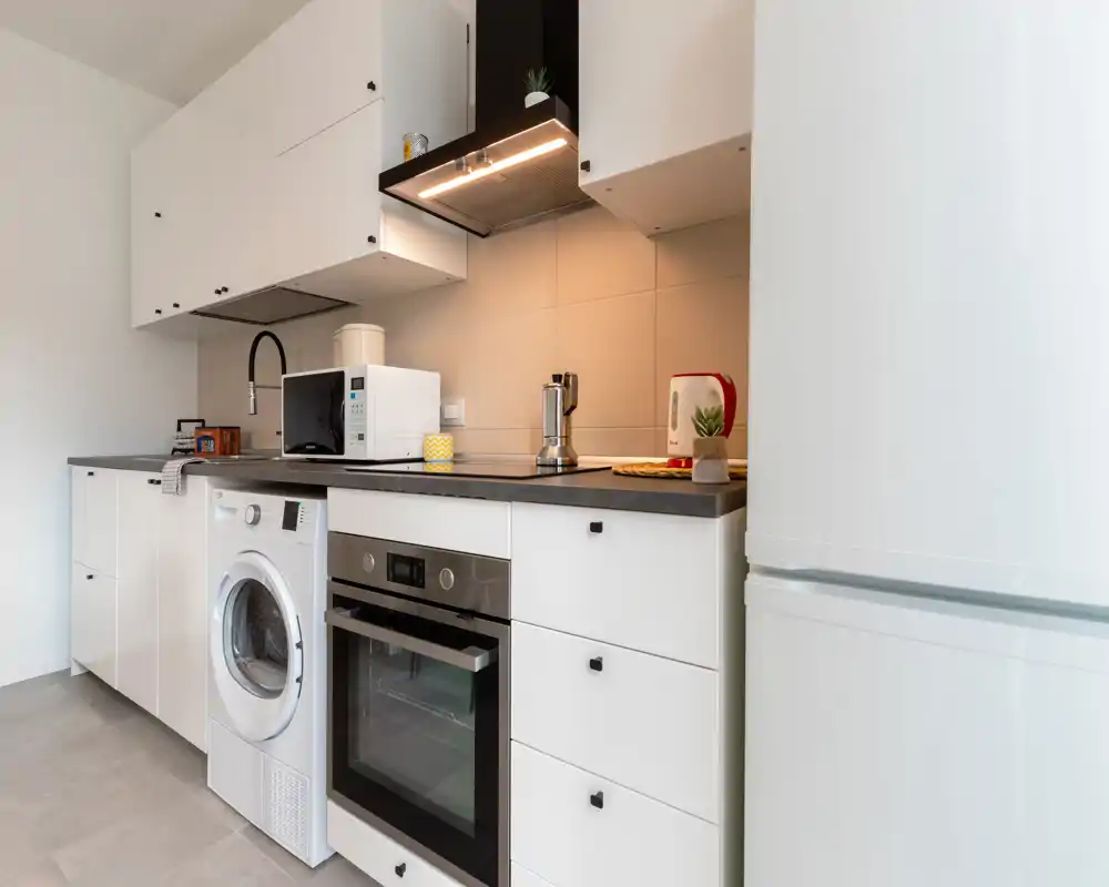 Cucinino moderno e luminoso con mobili bianchi, forno incassato, lavatrice e piano compatto con microonde e bollitore — perfetto per soggiorni in città efficienti.