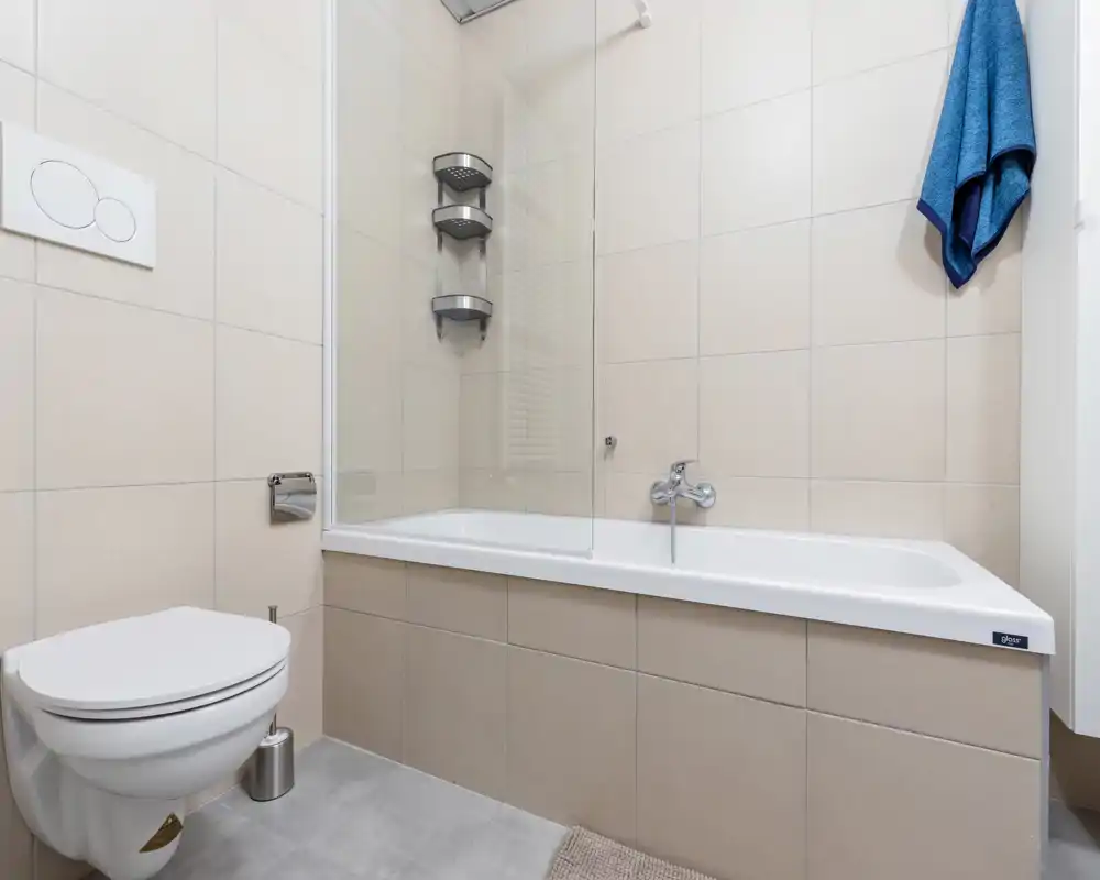 Bagno moderno e luminoso con vasca e wc sospeso, piastrelle beige neutre e parete doccia in vetro — pulito e ben curato.