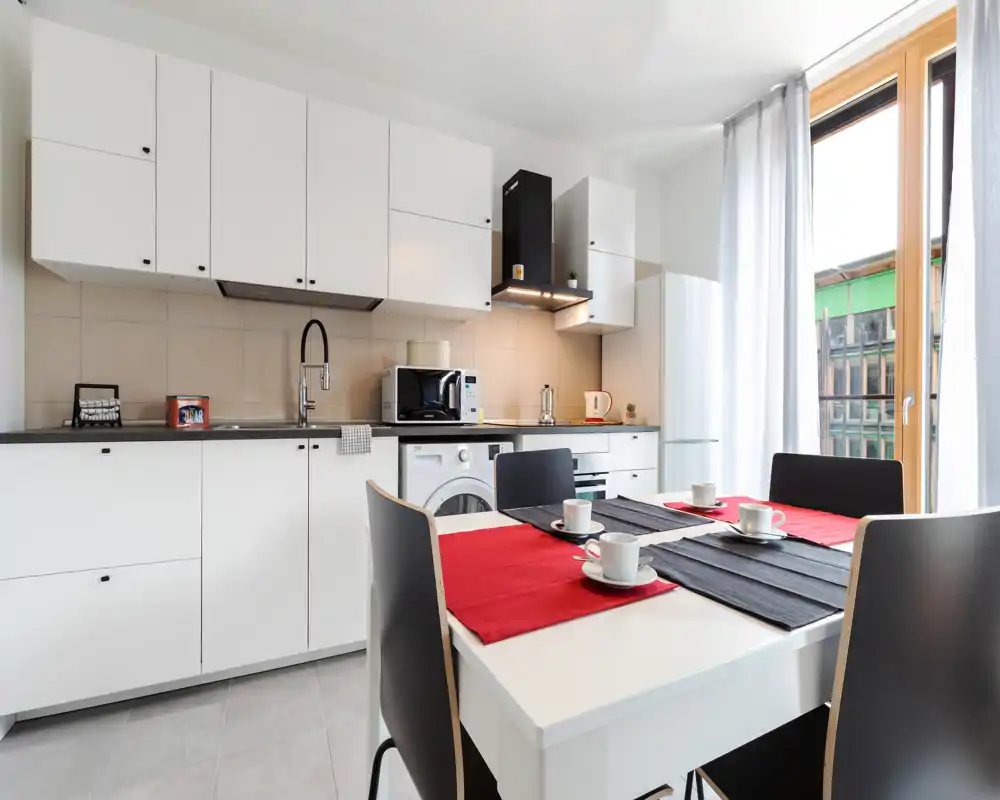 Cucina moderna e luminosa con mobili bianchi, elettrodomestici integrati e un tavolo per quattro vicino a una grande finestra che lascia entrare luce naturale.