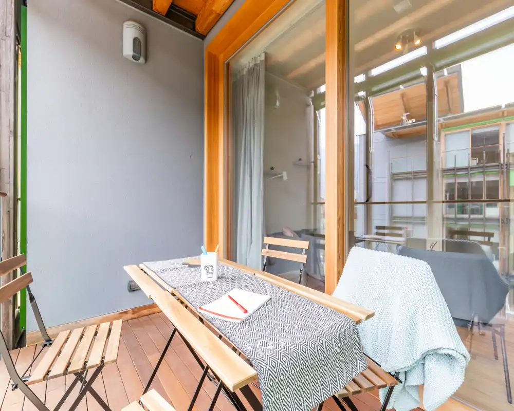 Balcone soleggiato arredato con tavolo in legno e sedie pieghevoli, tessili accoglienti e accesso diretto tramite grandi porte vetrate — ideale per il caffè del mattino.