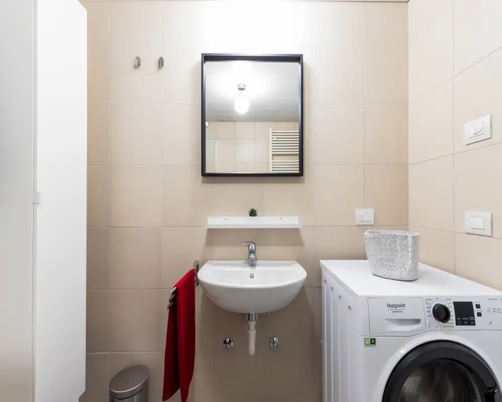 Bagno moderno e luminoso con lavabo a parete, specchio-scapoliera e lavatrice posizionata accanto al lavabo.