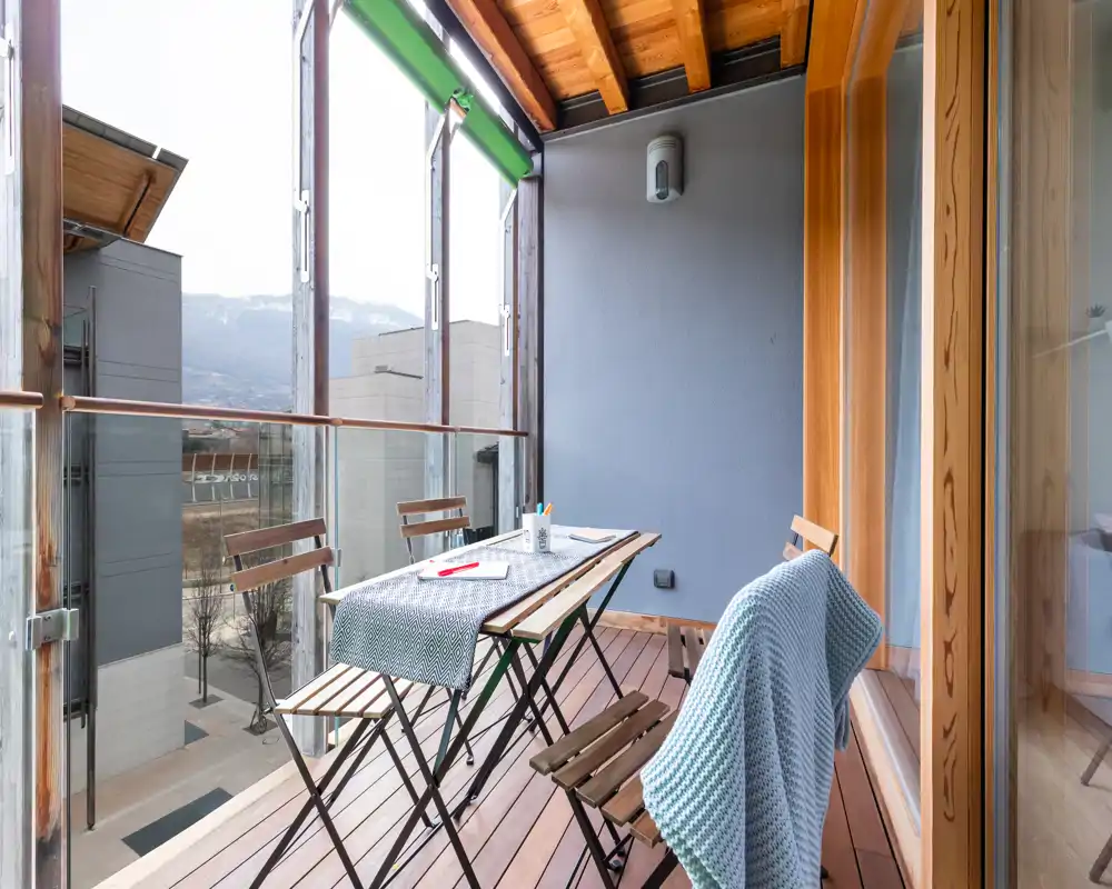 Balcone chiuso soleggiato con pavimento in legno e tavolo richiudibile con sedie — un angolo accogliente per caffè o pasti leggeri con vista su città e montagne.