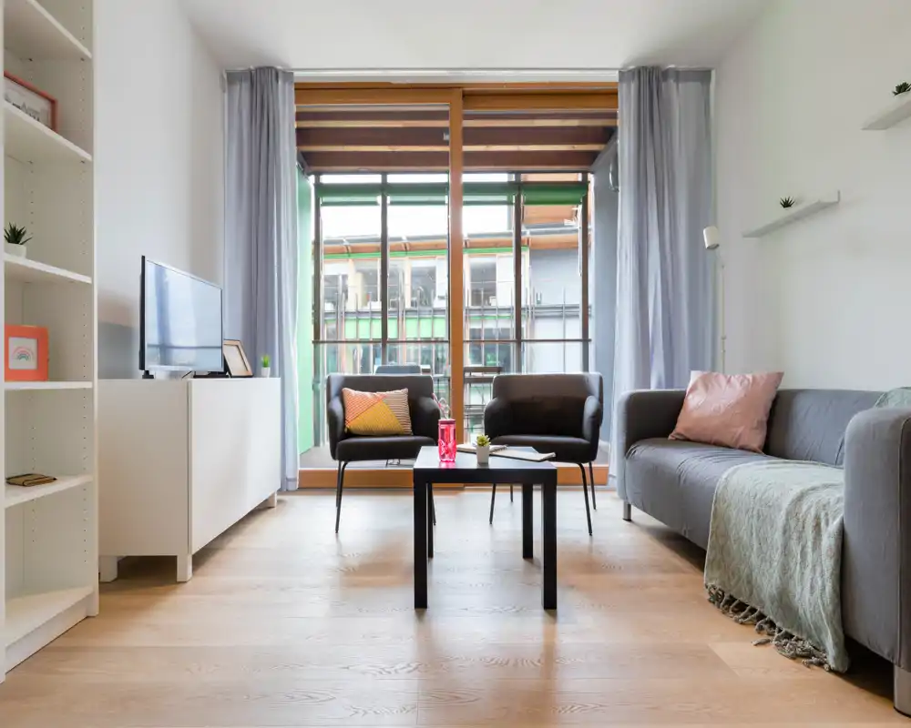 Soggiorno luminoso e moderno con ampia finestra, due poltrone e un divano comodo con cuscini e plaid. L’arredamento essenziale e la luce naturale rendono l’ambiente accogliente per il relax o gli ospiti.