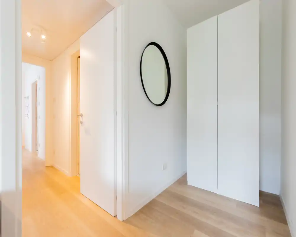 Ingresso luminoso e minimal con pavimento in legno chiaro, armadio bianco e specchio rotondo: un primo impatto pulito e accogliente.