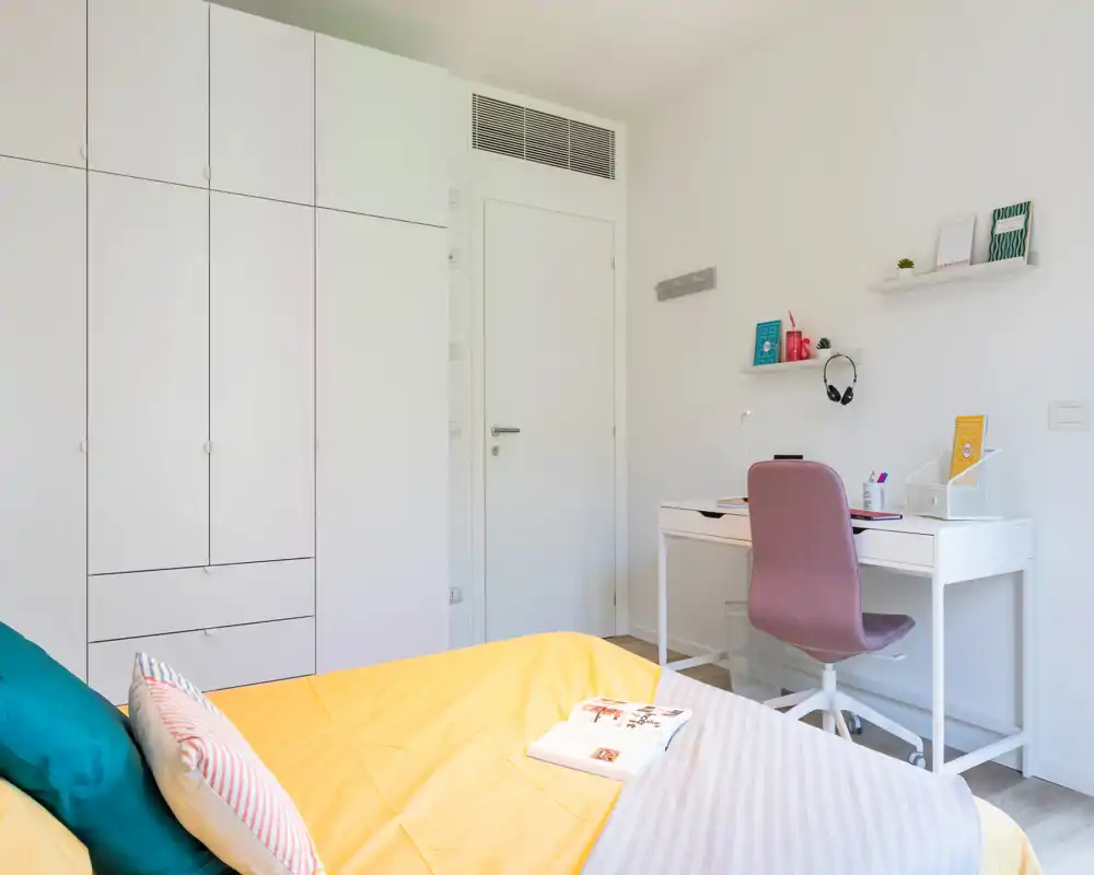 Dormitorio moderno y luminoso con cama ordenada, armarios empotrados blancos y un escritorio compacto con silla rosa — ideal para una persona o estudiante.