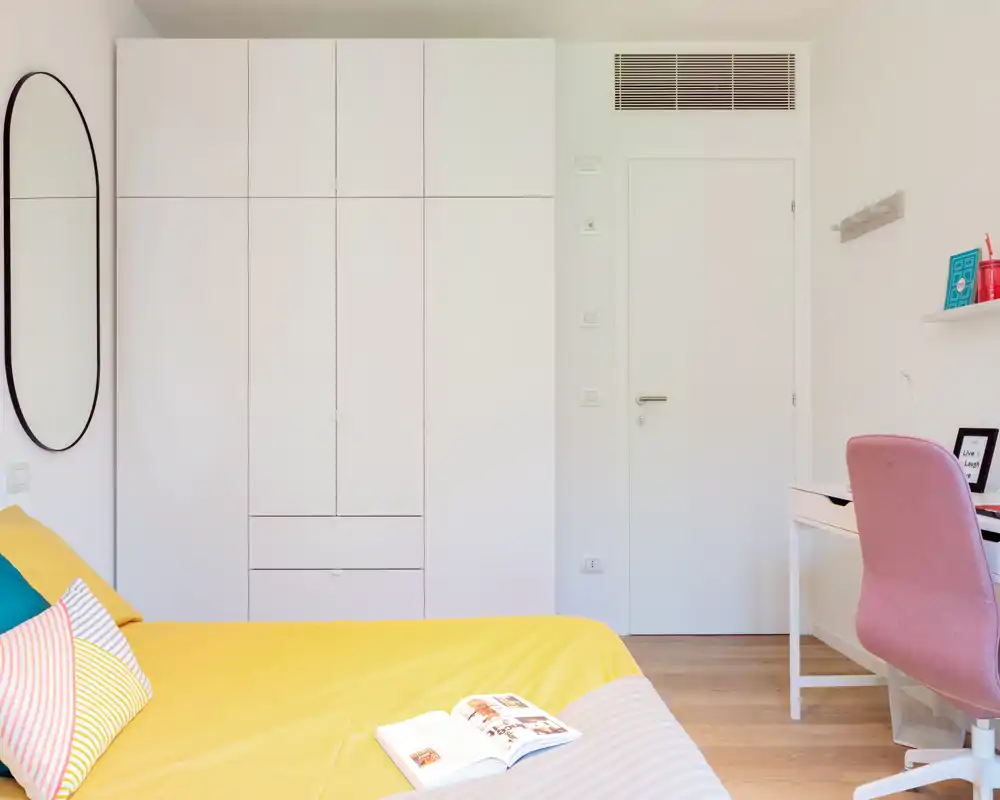 Dormitorio moderno y luminoso con colcha amarilla, cojines de colores y armario empotrado blanco. Espacio ordenado y acogedor, perfecto para anuncios de alquiler.