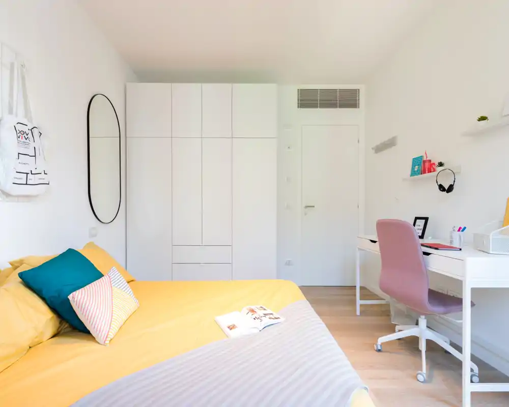 Dormitorio moderno y luminoso con cama grande vestida de amarillo, armario y escritorio ordenado. La luz natural y los cojines coloridos crean un ambiente fresco y acogedor, perfecto para un anuncio de alquiler.