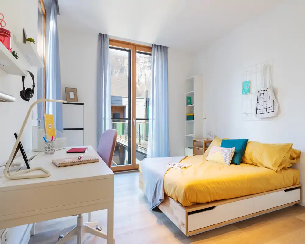 Dormitorio moderno y luminoso con cama individual de ropa de cama amarilla, escritorio junto a la ventana y grandes puertas acristaladas que aportan mucha luz natural.