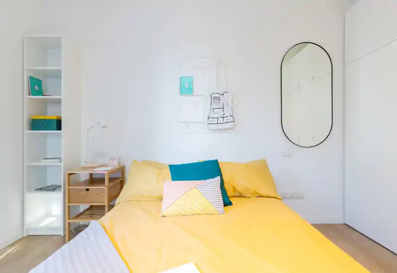 Dormitorio luminoso y moderno con gran colcha amarilla, cojines decorativos y paredes blancas; estantería minimalista y mesita completan el ambiente acogedor.