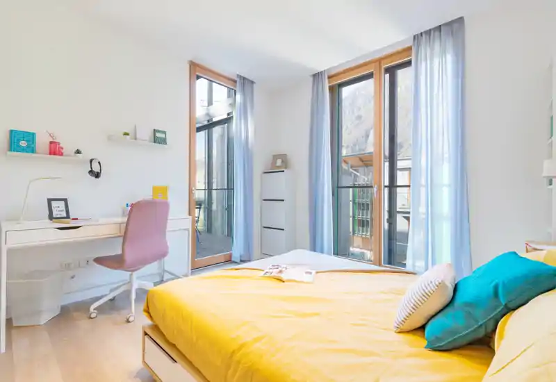 Dormitorio moderno y luminoso con cama completa con ropa de cama amarilla, grandes ventanas de suelo a techo y un escritorio compacto: ideal para luz natural y un rincón de trabajo acogedor.