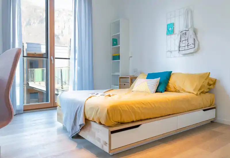 Dormitorio moderno y luminoso con cama con almacenamiento, ropa de cama amarilla, cojines decorativos y luz natural desde una gran ventana-balcon.