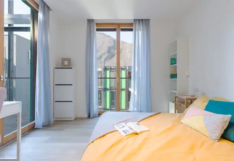 Dormitorio moderno y luminoso con una gran ventana y acceso al balcón. La cama doble está bien hecha con ropa de cama colorida, suelo de madera clara y estanterías sencillas para almacenamiento.