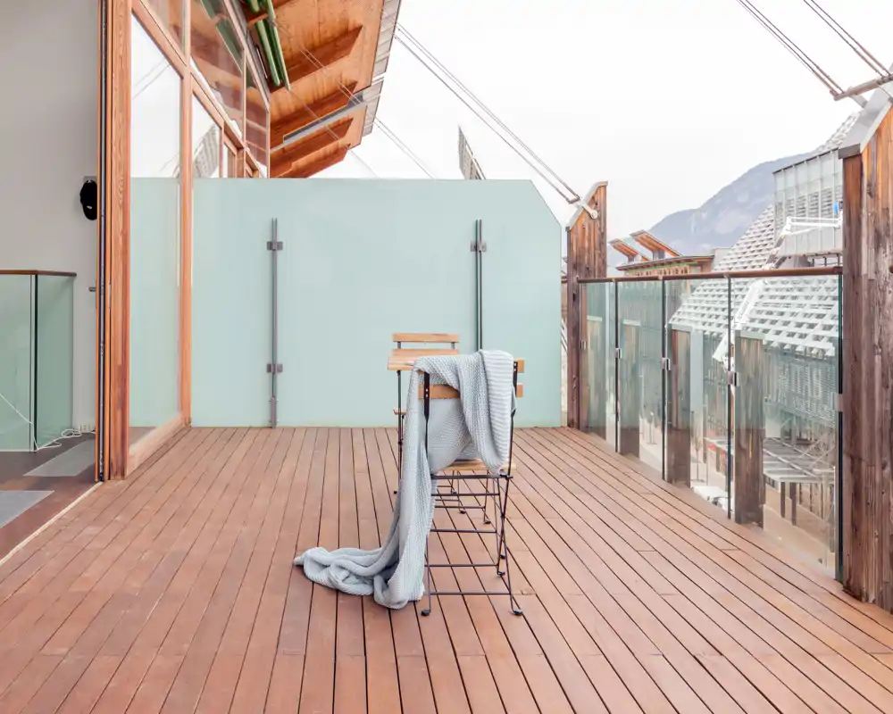 Sunny wooden terrace with modern glass balustrade and a small chair draped with a cozy blanket — bright, minimal outdoor space with mountain views.