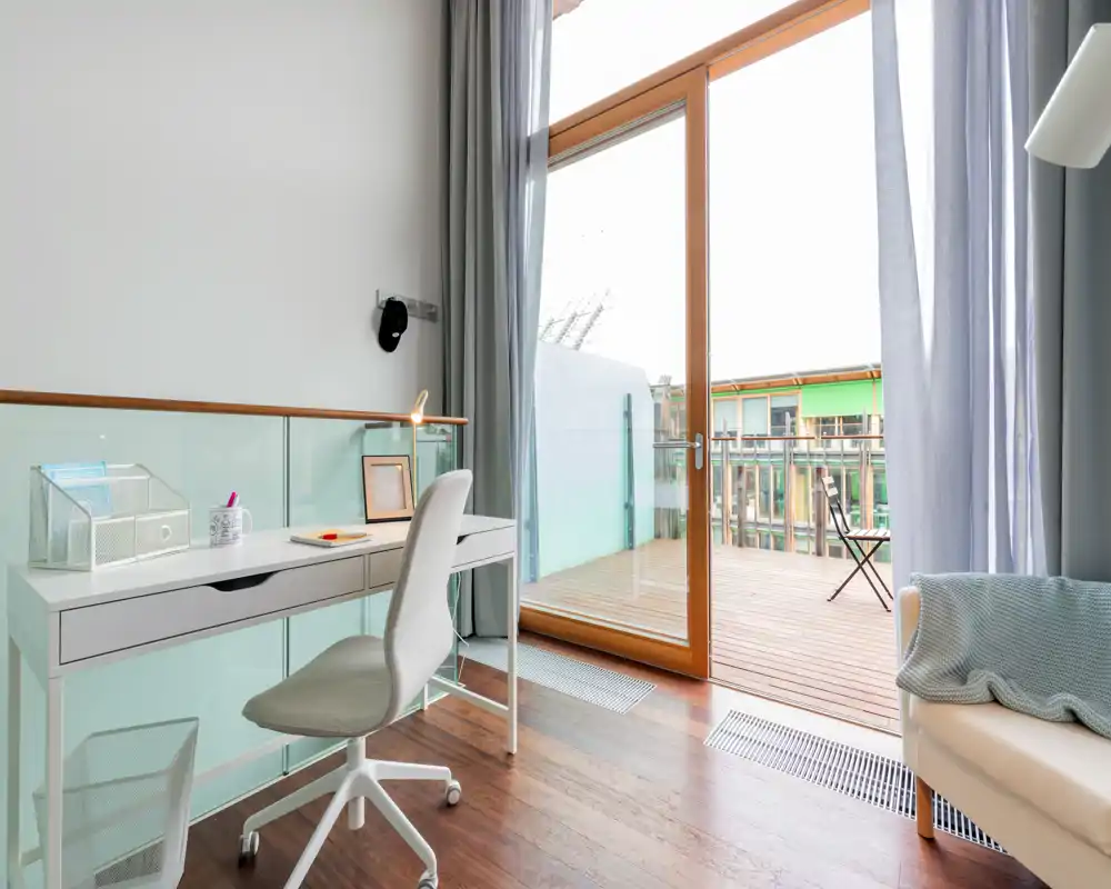Bright, modern room featuring a compact white desk and ergonomic chair beside a large sliding glass door that opens onto a wooden balcony — ideal as a small home office with outdoor access.