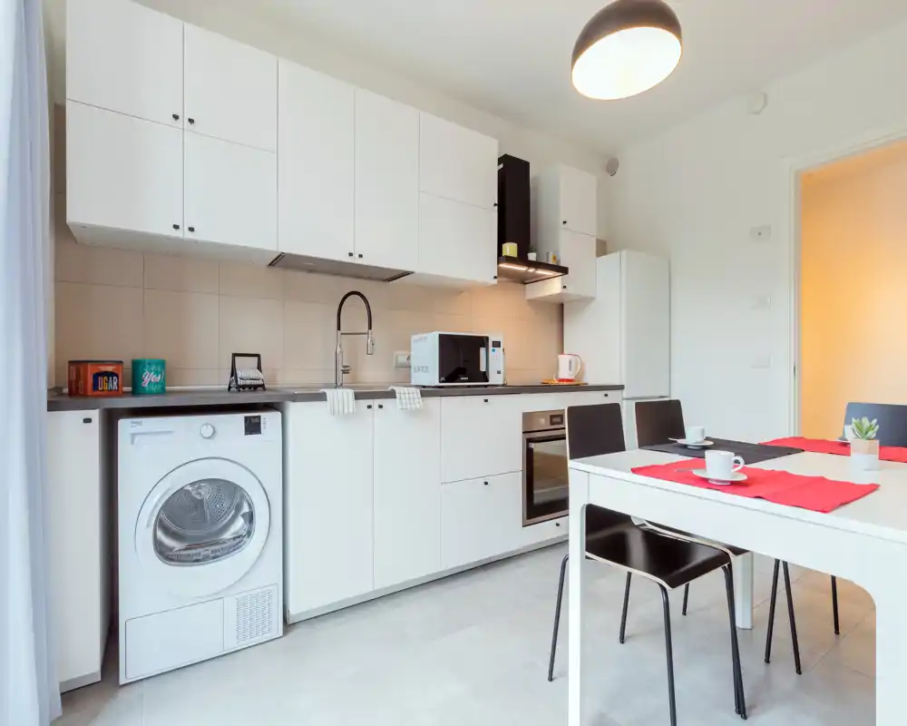 Bright modern kitchen with white cabinets, integrated oven, microwave and a washing machine under the counter; a small dining table with black chairs is set for coffee.