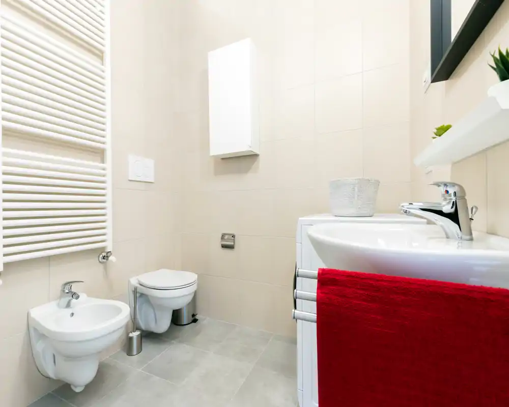 Modern, bright bathroom with wall-mounted toilet and bidet, white sink with chrome faucet and a red towel. Neutral tiles and a heated towel rail create a clean, contemporary look.