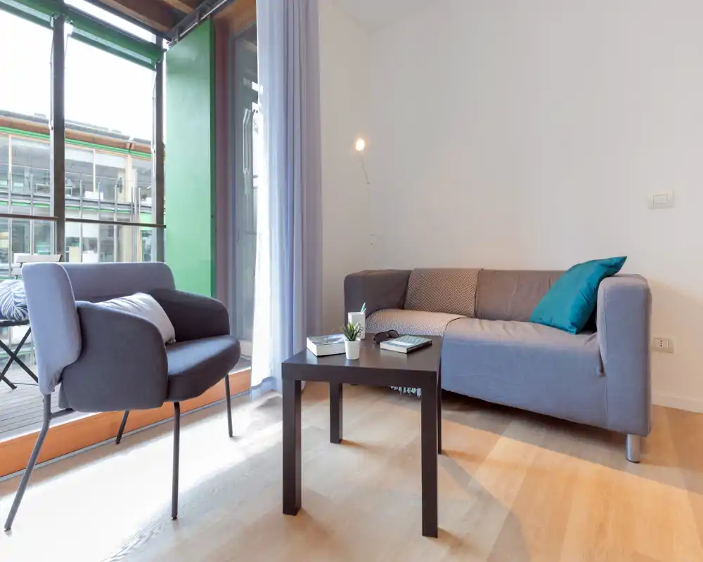 Bright, modern living area with a compact grey sofa, armchair and coffee table by a large window leading to a balcony — ideal for a cozy sitting space with natural light.