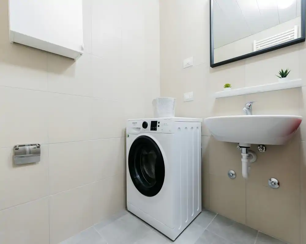 Compact, modern bathroom corner with a front-loading washing machine, wall-mounted sink and mirror. Neutral tile walls and simple fixtures create a clean, functional space.