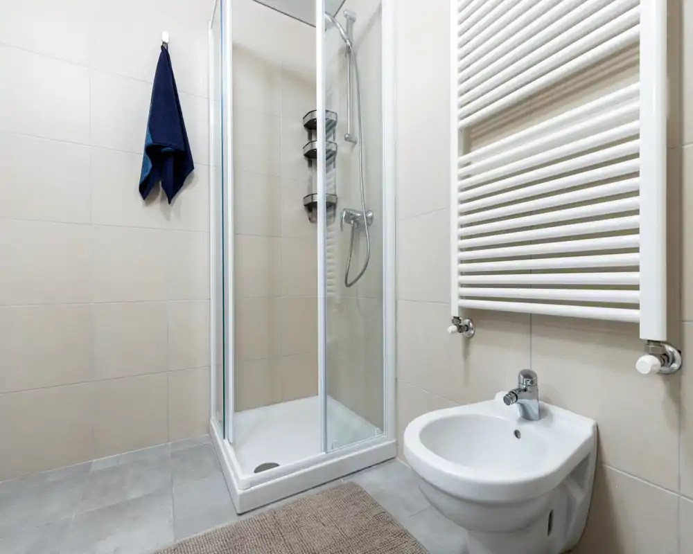 Compact modern bathroom with a glass shower enclosure, wall-mounted bidet, heated towel rail and neutral tile finishes.
