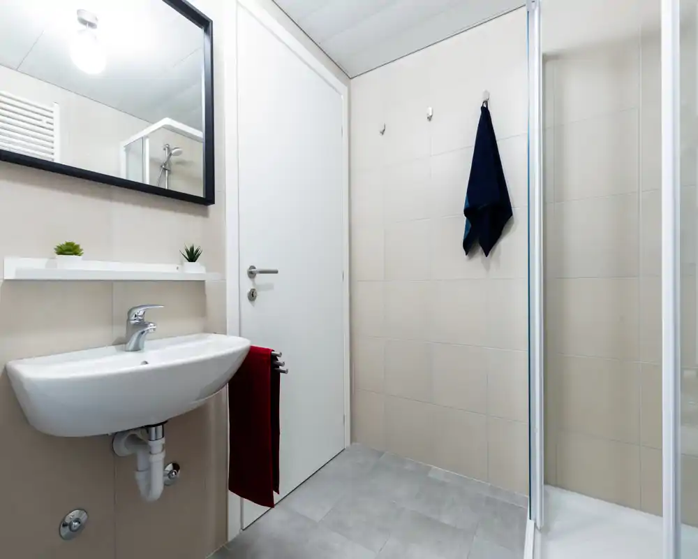 Bright, modern bathroom with a wall-mounted sink, large mirror, glass-enclosed shower and simple hooks for towels — clean and well-lit.