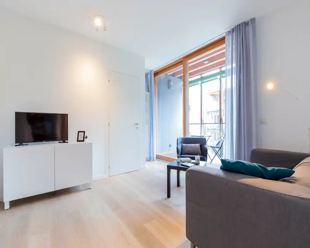 Bright, modern living room with a comfortable sofa, armchair and coffee table opening onto a small balcony. Large window floods the space with natural light; minimalist decor and soft tones create a calm, welcoming atmosphere.