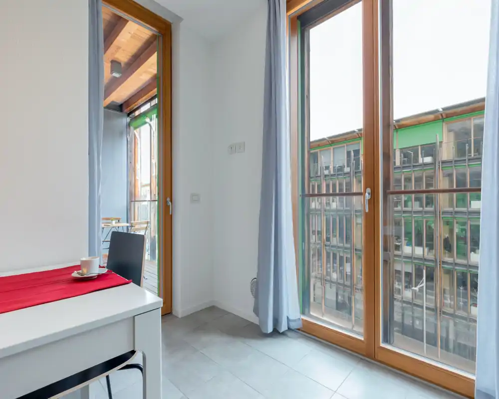 Bright small kitchen/dining corner with large floor-to-ceiling windows and a compact table set with a chair and cup; natural light and a balcony just outside provide an airy feel.