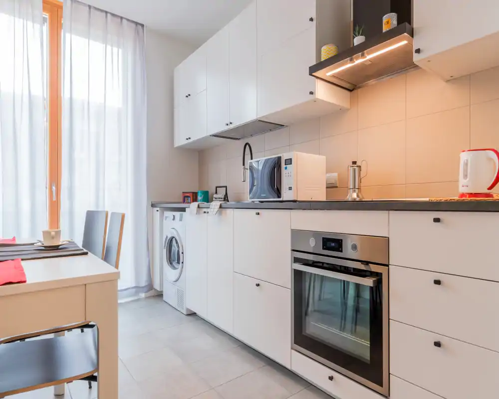 Bright, modern kitchen featuring white cabinets, integrated oven, microwave and a washing machine next to a dining table; large window with sheer curtains provides soft natural light.