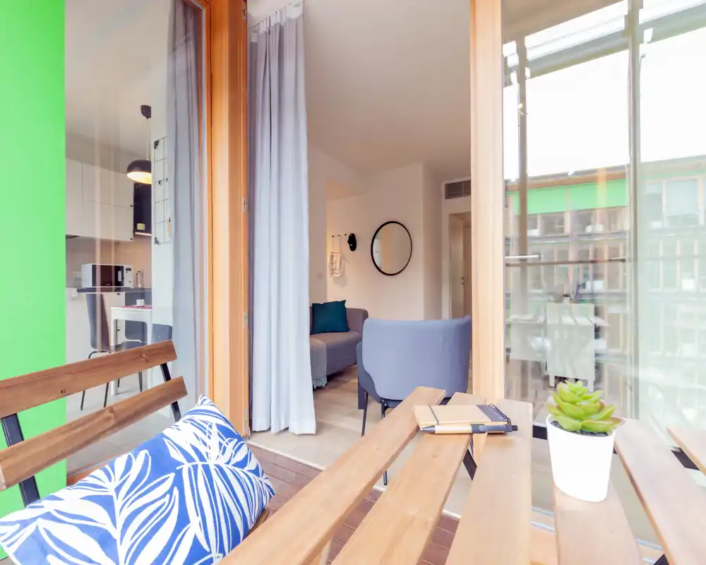 Sunny living area opening onto a small balcony: comfortable seating, decorative mirror and soft textiles, with large sliding doors letting in ample natural light.