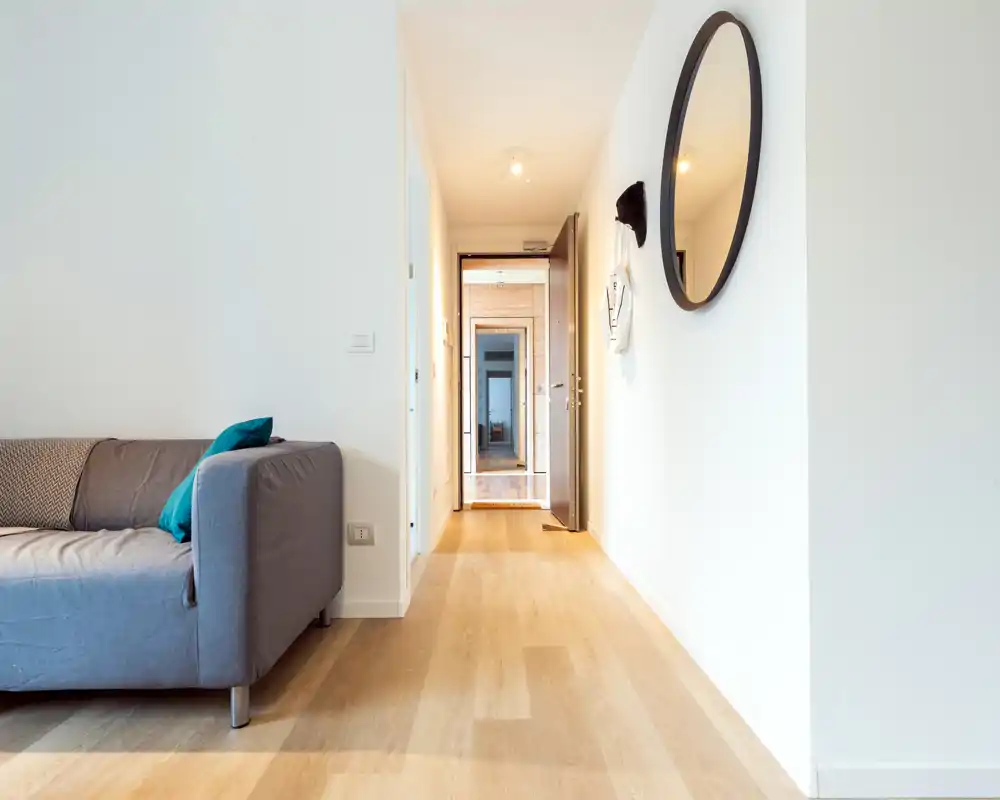 Bright, modern living area seen from the hallway with a grey sofa and accent cushions; clean lines and light wood flooring create an airy, contemporary feel.