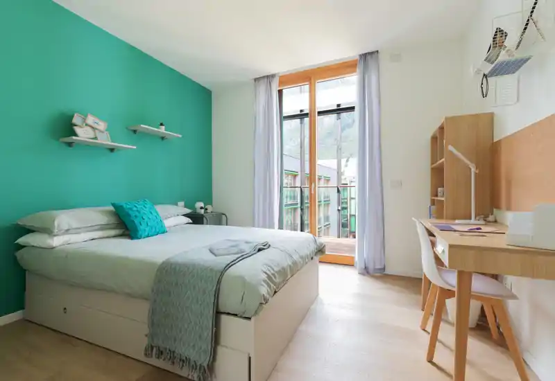 Bright, modern bedroom with a double bed against a teal accent wall, soft linens, a study desk by the window and a balcony providing natural light.
