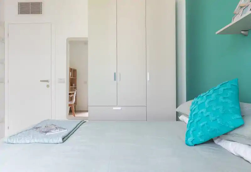 Bright, modern bedroom with a neatly made bed, teal accent wall and built-in wardrobe; crisp linens and decorative cushion create a calm, inviting atmosphere.