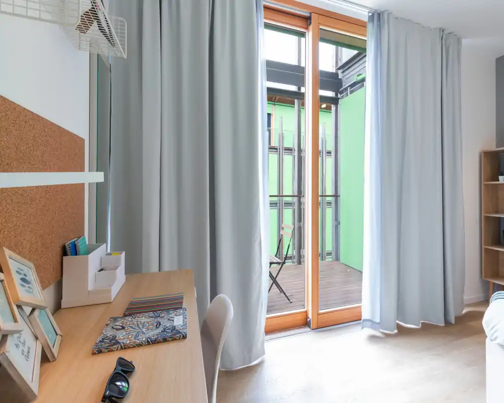 Bright bedroom corner with a wooden desk, study chair and light curtains opening onto a small balcony. The bed is only partially visible at the right edge; the image highlights the workspace and natural light.