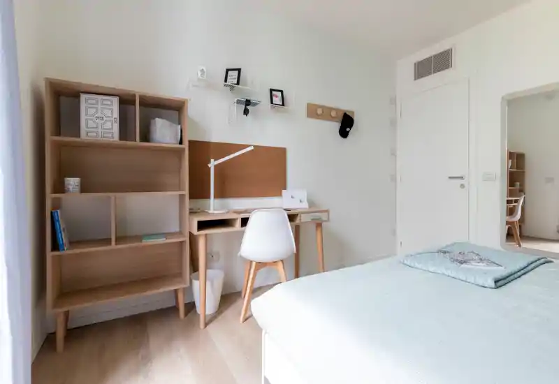 Bright, minimalist bedroom with a neatly made bed and a wooden desk workspace. Clean Scandinavian decor, natural light, and practical shelving create a calm study-and-sleep area ideal for a student or professional.
