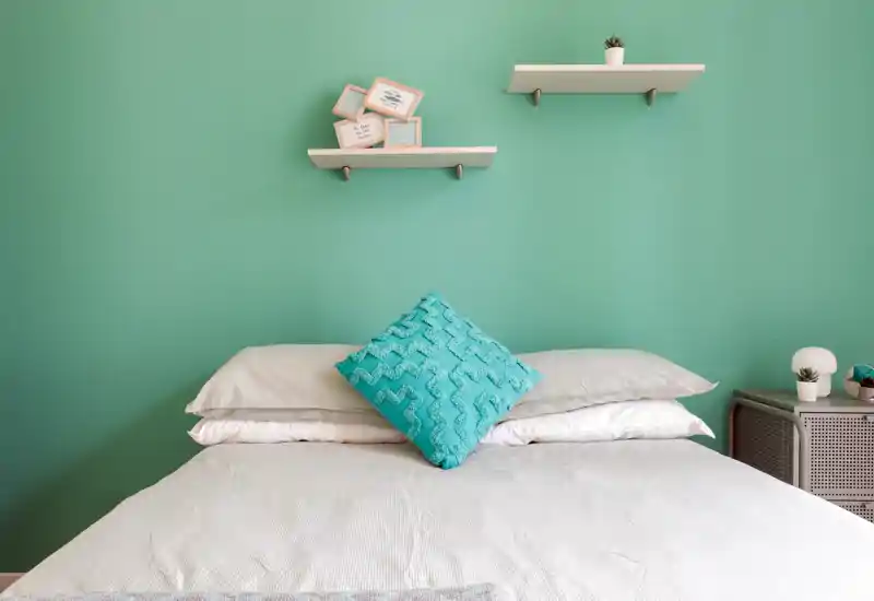 Bright, stylish bedroom with a centered bed, layered pillows and a turquoise accent cushion against a soft green wall with minimalist shelves.