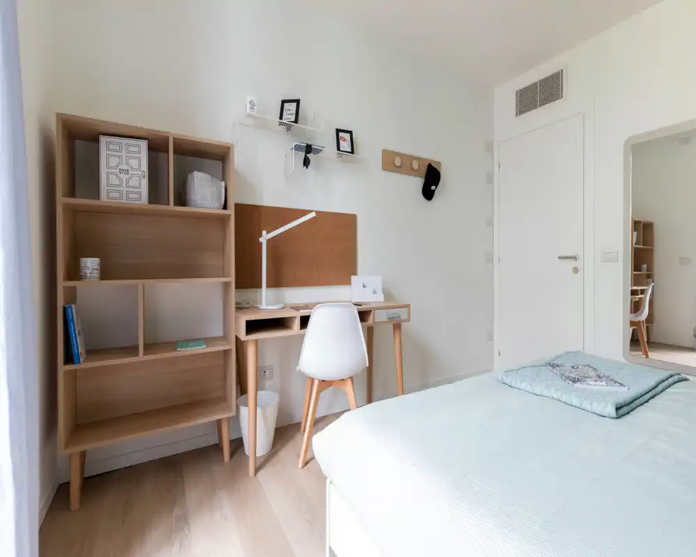 Camera da letto luminosa e moderna con angolo letto visibile, scrivania in legno e scaffali — ideale per studenti o smart worker.
