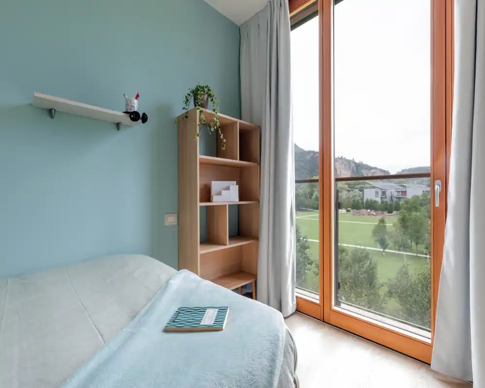 Bright, modern single bedroom with a neatly made bed, pastel blue wall, open shelving and a large window with a scenic view. Soft textiles and natural light make it inviting for short-term rentals.