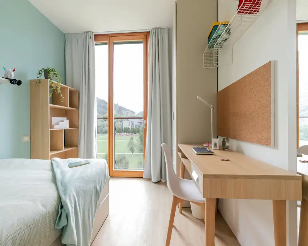 Bright, minimal bedroom with a comfortable bed, wooden desk and a large window overlooking green scenery. The room features soft pastel tones, light wood furniture and practical storage for a calm, study-friendly atmosphere.