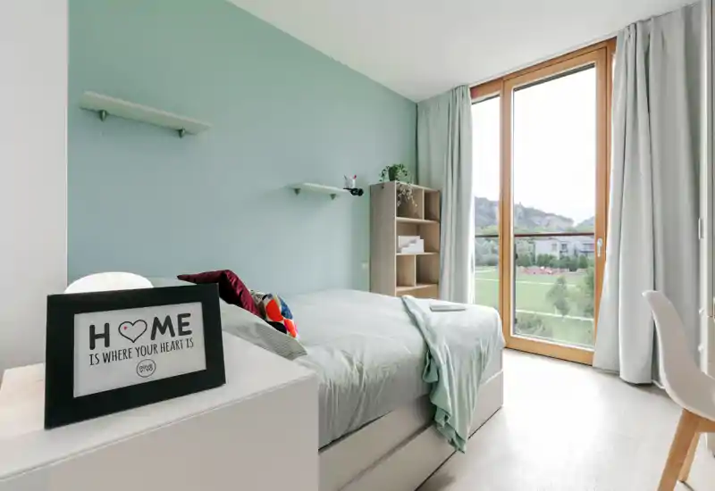 Bright, modern bedroom with a neatly made bed, soft green accent wall and large floor-to-ceiling window overlooking green space — a calm, well-styled private room ideal for relaxing.