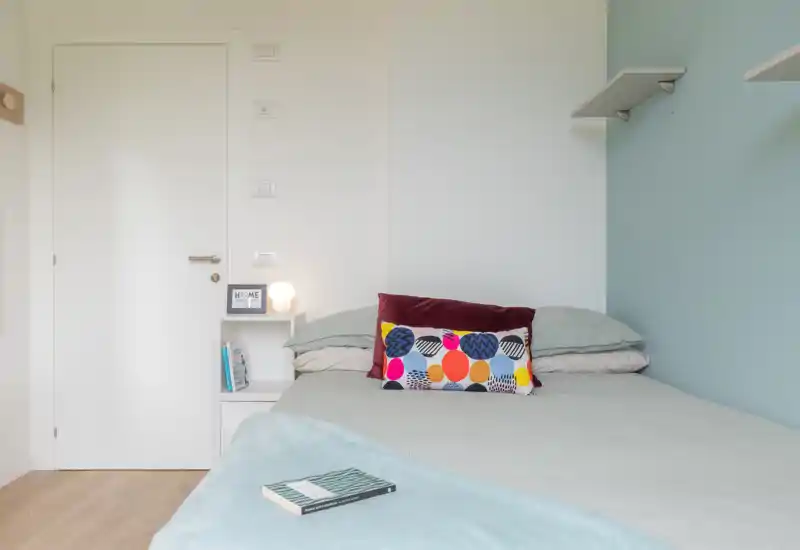 Bright, minimalist bedroom with a neatly made double bed, colorful decorative cushion and a small bedside shelf with lamp — calm, modern sleeping space.