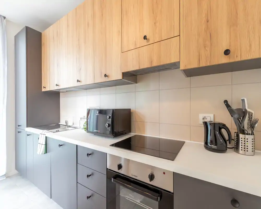 Modern compact kitchen with wood upper cabinets, matte lower cabinets, induction cooktop, oven and small appliances on a clean white countertop. Bright, well-styled space ideal for rental listings.