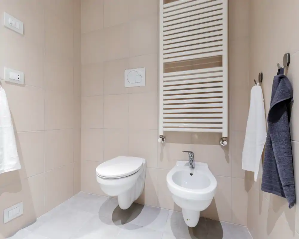Clean, modern bathroom with wall-mounted toilet and bidet, neutral tiles and a heated towel radiator. Bright, minimal and well-lit space suitable for rental listings.