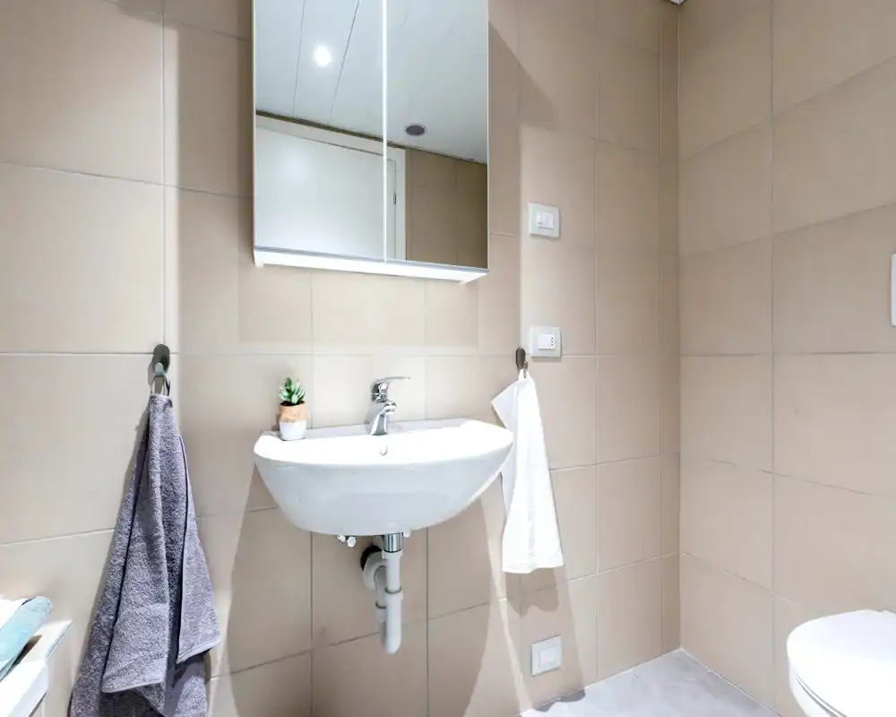 Clean, modern bathroom with a wall-mounted sink, mirrored cabinet and neutral beige tiles. Bright, well-maintained space suitable for rental listings.